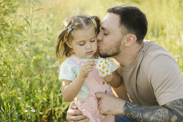 Father kissing daughter holding daisies by plants - SEAF00196