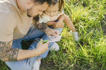 Neugieriges Mädchen sitzt bei ihrem Vater und schneidet übertragbare Tattoos auf Gras - SEAF00202