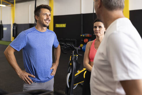 Lächelnder Sportler mit Hand auf der Hüfte im Gespräch mit Freunden im Fitnessstudio - JPTF00925