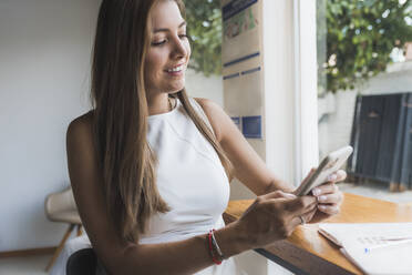 Lächelnde Frau, die ein Smartphone benutzt, sitzt am Tisch in einem Café - DSIF00582