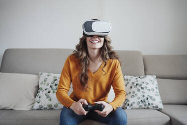 Woman playing video game wearing virtual reality headset at home - EBBF04938