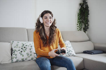 Happy woman with headphones playing game through joystick at home - EBBF04940