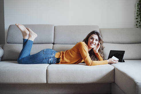 Junge Frau mit Kreditkarte und Tablet-PC zu Hause auf dem Sofa liegend - EBBF04954