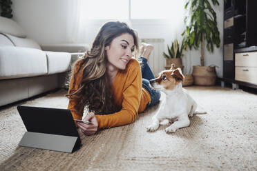 Young woman with tablet PC looking at pet dog lying on floor - EBBF04959