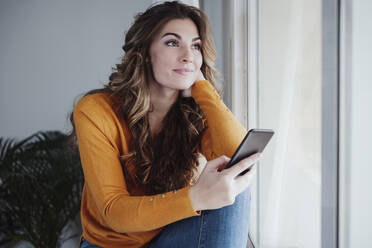 Contemplating woman with smart phone sitting near window at home - EBBF04970