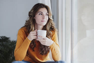 Junge Frau hält Kaffeetasse am Fenster zu Hause - EBBF04971