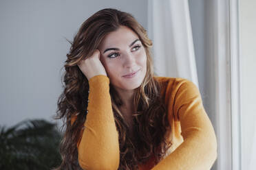 Thoughtful woman with head in hand sitting near window at home - EBBF04977