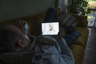 Doctor talking with patient lying on sofa through video call at home - UUF25241