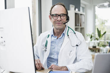 Lächelnder Arzt vor einem Computermonitor in einer Klinik - UUF25249