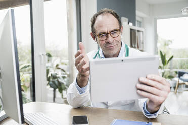 Lächelnder Arzt mit digitalem Tablet gestikuliert bei einem Videogespräch in einer Klinik - UUF25259