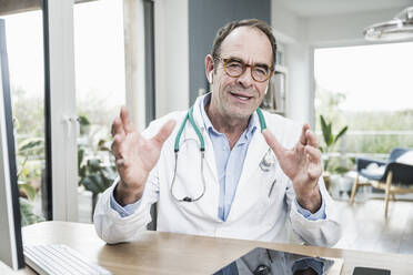 Doctor wearing eyeglasses gesturing at desk in hospital - UUF25260