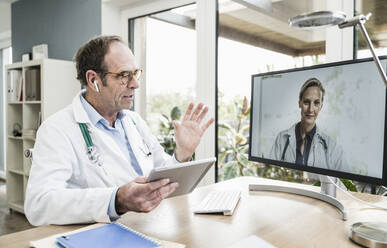 Doctor with digital tablet discussing with colleague on video call through computer at office - UUF25263