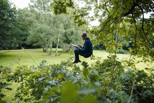 Geschäftsmann, der mit einem Seil an einem Baum hängt und ein digitales Tablet im Park benutzt - GUSF06522