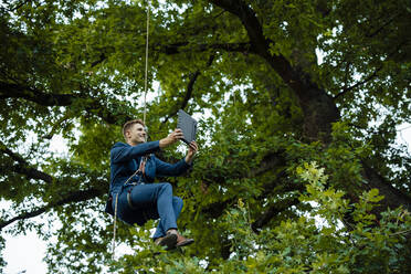 Smiling businessman tied up with rope on tree doing video call through tablet PC - GUSF06525