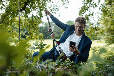 Smiling businessman using smart phone hanging on rope in park - GUSF06528