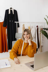 Junge Geschäftsfrau mit Hand am Kinn sitzt am Schreibtisch in einem kreativen Büro - TCEF02119