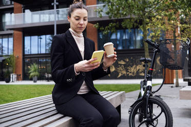 Businesswoman with disposable cup using mobile phone on bench by bicycle - ASGF01831