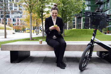 Smiling businesswoman using smart phone sitting on bench - ASGF01833