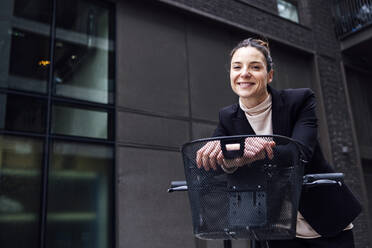 Smiling businesswoman leaning on electric bicycle with basket - ASGF01839