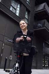 Smiling businesswoman leaning on electric bicycle with basket in front of office building - ASGF01840