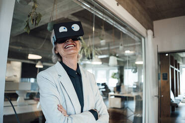 Smiling businesswoman with arms crossed using virtual reality headset in office - JOSEF05990