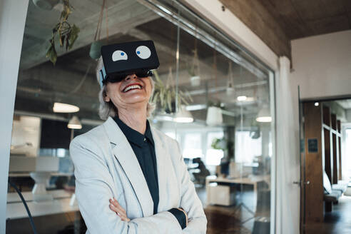 Lächelnde Geschäftsfrau mit verschränkten Armen, die ein Virtual-Reality-Headset im Büro benutzt - JOSEF05990