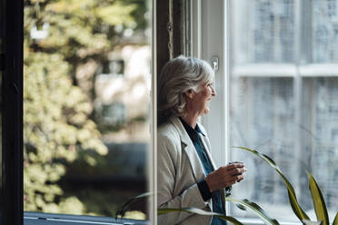 Senior businesswoman with coffee cup looking out of office window - JOSEF05993