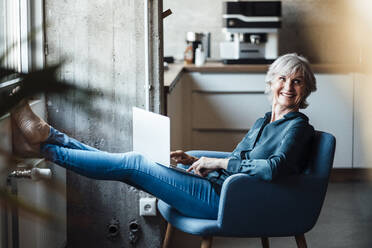 Businesswoman sitting with laptop on armchair in office - JOSEF05995