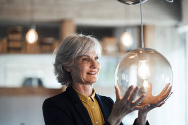 Smiling businesswoman looking at illuminated pendant light in office - JOSEF06012