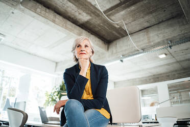 Businesswoman with hand on chin sitting on desk - JOSEF06023