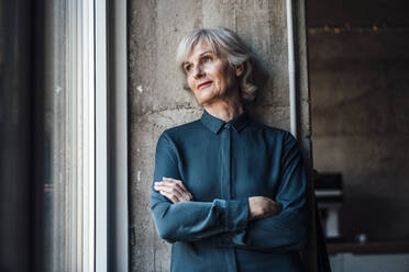 Thoughtful businesswoman with arms crossed at office window - JOSEF06058