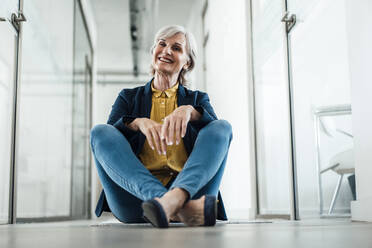 Smiling businesswoman sitting with cross-legged at office corridor - JOSEF06078