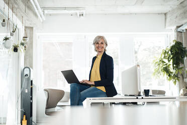 Senior businesswoman with laptop sitting on office desk - JOSEF06088