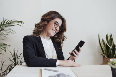 Smiling businesswoman with eyeglasses using smart phone at office - EBBF05026