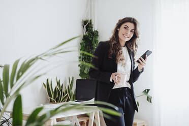 Businesswoman with coffee mug holding smart phone near desk at office - EBBF05038