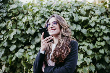 Lächelnde Geschäftsfrau, die vor einer Hecke eine Sprachnachricht über ihr Smartphone verschickt - EBBF05071