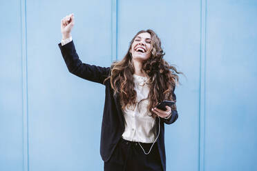 Carefree businesswoman enjoying music on smart phone in front of blue wall - EBBF05074