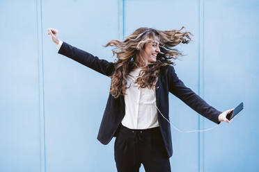 Happy businesswoman dancing holding smart phone in front of blue wall - EBBF05075