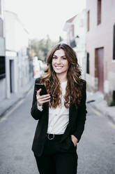 Young businesswoman listening music through smart phone standing in alley - EBBF05078