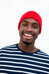 Cheerful bearded African American man in red hat looking at camera while standing on white background on street - ADSF32437