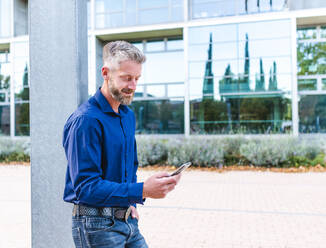 Adult male with gray hair and beard standing with hand in pocket of jeans near pillar and browsing mobile phone - ADSF32446