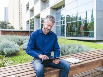 Positiver erwachsener männlicher Unternehmer, der während einer Geschäftsreise auf einer Holzbank neben einem Netbook an einem Tablet arbeitet - ADSF32448