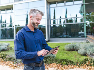 Konzentrierter bärtiger Mann in Hemd und Jeans, der auf seinem Tablet scrollt, während er aus der Ferne in einem Park in der Nähe eines modernen Bürogebäudes Arbeitsaufgaben erledigt - ADSF32449