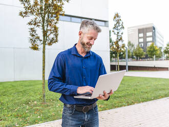 Positiver Mann, der im Park steht und auf einem modernen Netbook tippt, während er ein Online-Geschäftsprojekt durchführt - ADSF32450