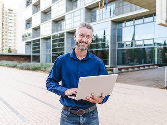 Positiver Mann, der im Park steht und auf einem modernen Netbook tippt, während er ein Online-Geschäftsprojekt durchführt - ADSF32451