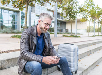 Positiv, männlich, der sein Smartphone benutzt, während er auf einer Steintreppe in der Nähe seines Koffers auf einer Geschäftsreise sitzt - ADSF32452