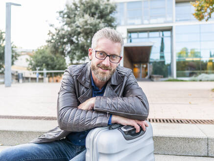 Positive bearded man folding hands on suitcase while sitting on stone steps during work trip and looking at camera - ADSF32453