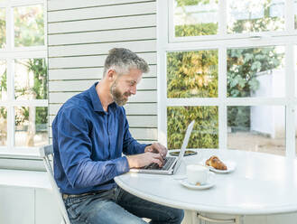 Männlicher Unternehmer bei der Telearbeit mit einem Netbook am Tisch mit Smartphone und Kaffee mit Croissant in einem Straßencafé - ADSF32457