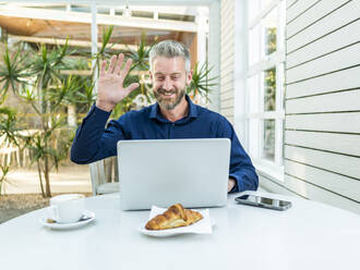 Smiling male entrepreneur waving hand to camera of netbook during online video chat at table with smartphone and coffee with croissant in outdoor cafe - ADSF32458