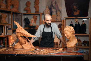 Pensive bearded male woodworker in apron standing at table with carved wooden sculptures and professional instruments during work in joinery - ADSF32459